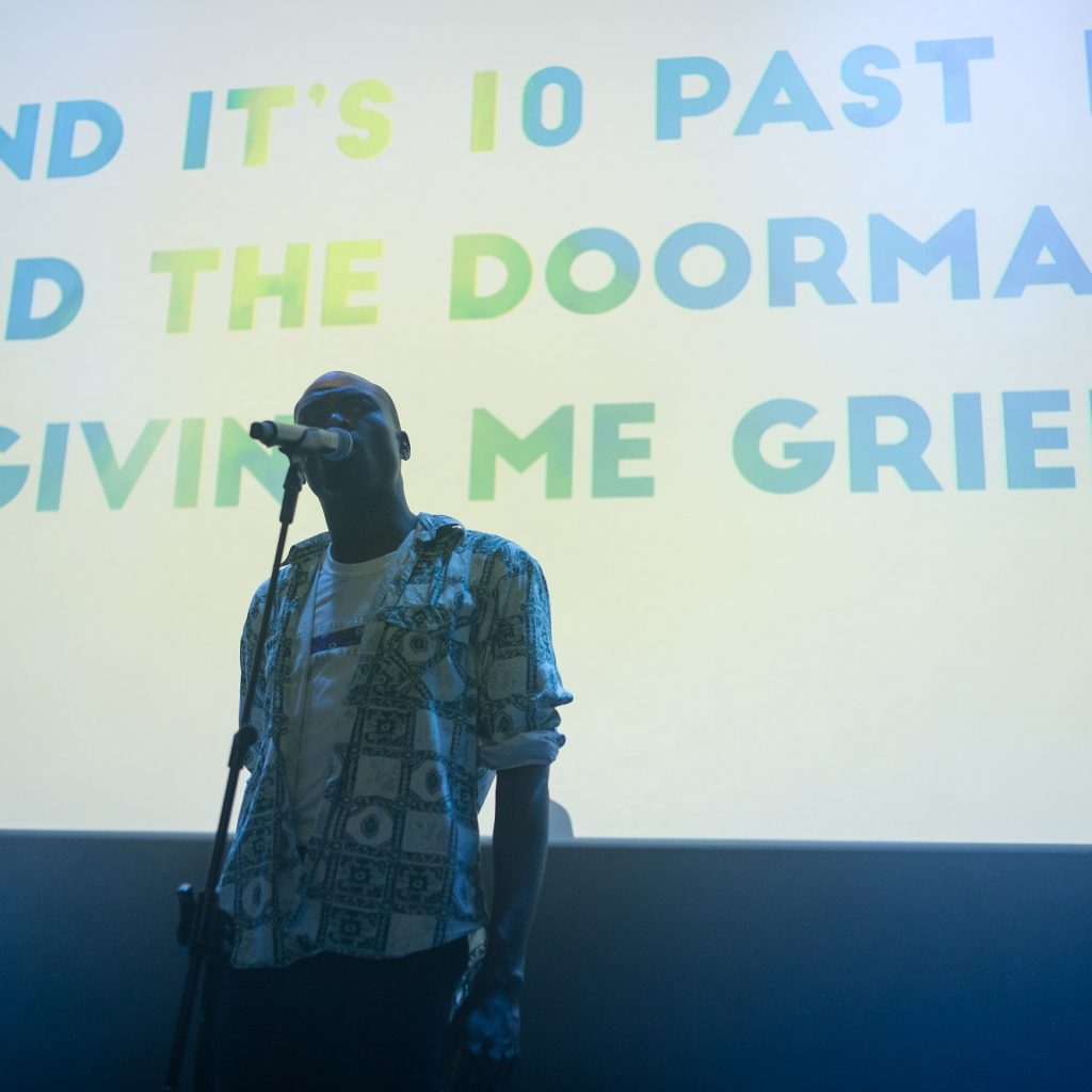 A young, black man stands on stage in front of a microphone with a screen behind with writing on it.