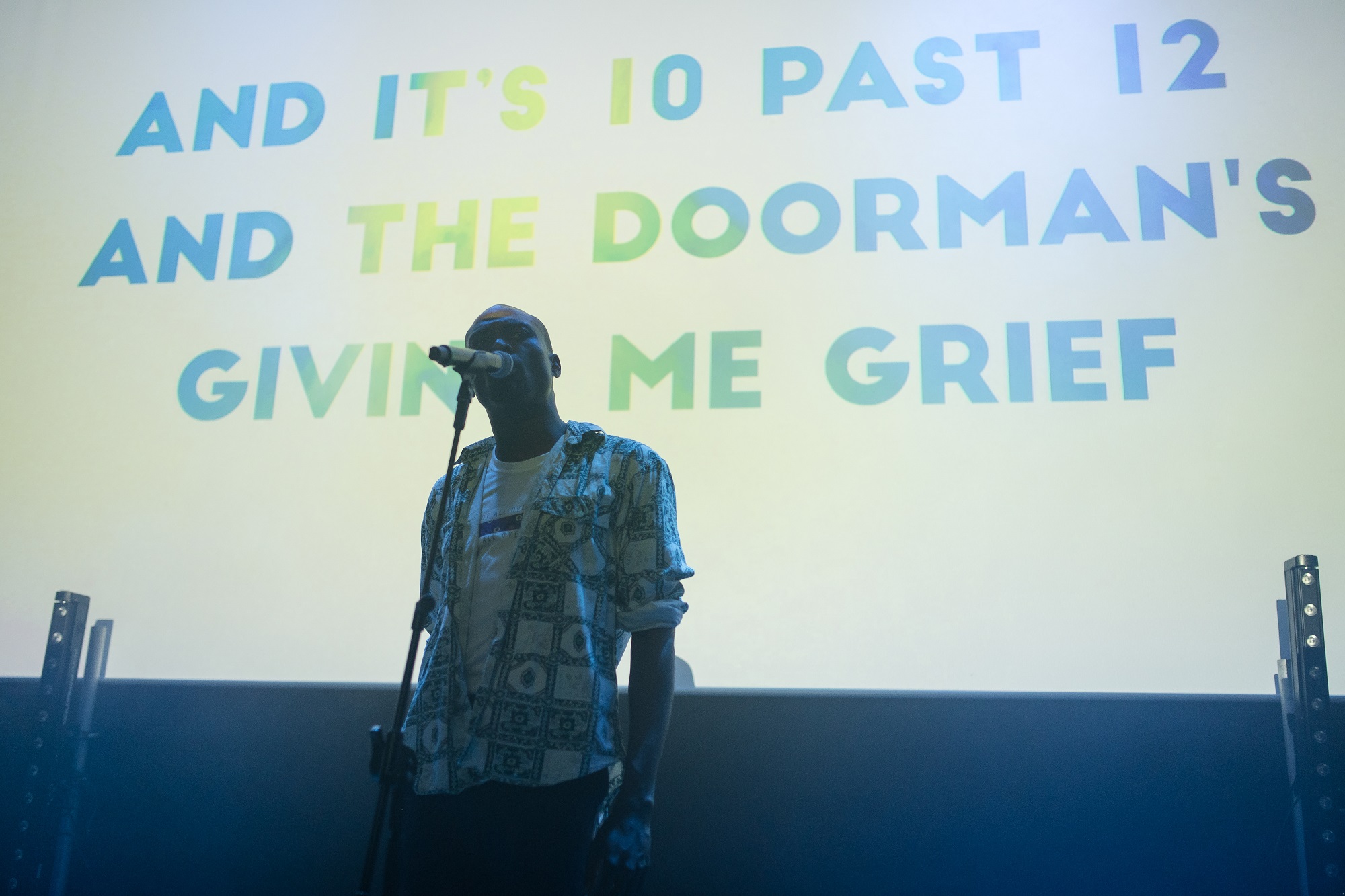 A young, black man stands on stage in front of a microphone with a screen behind with writing on it.
