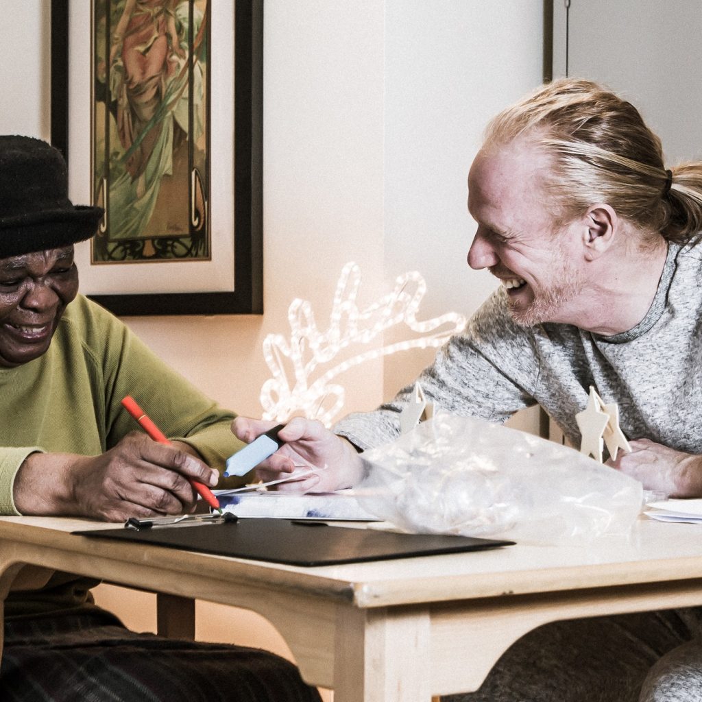 An older black woman and a younger white man seated at a table, doing craft and laughing.