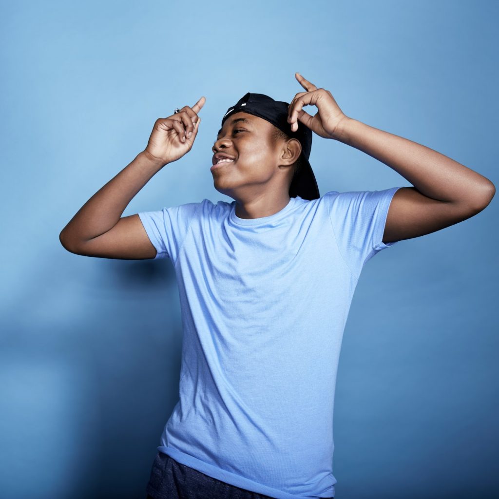 A young, black man in a blue T-shirt and a cap dances with his arms up.