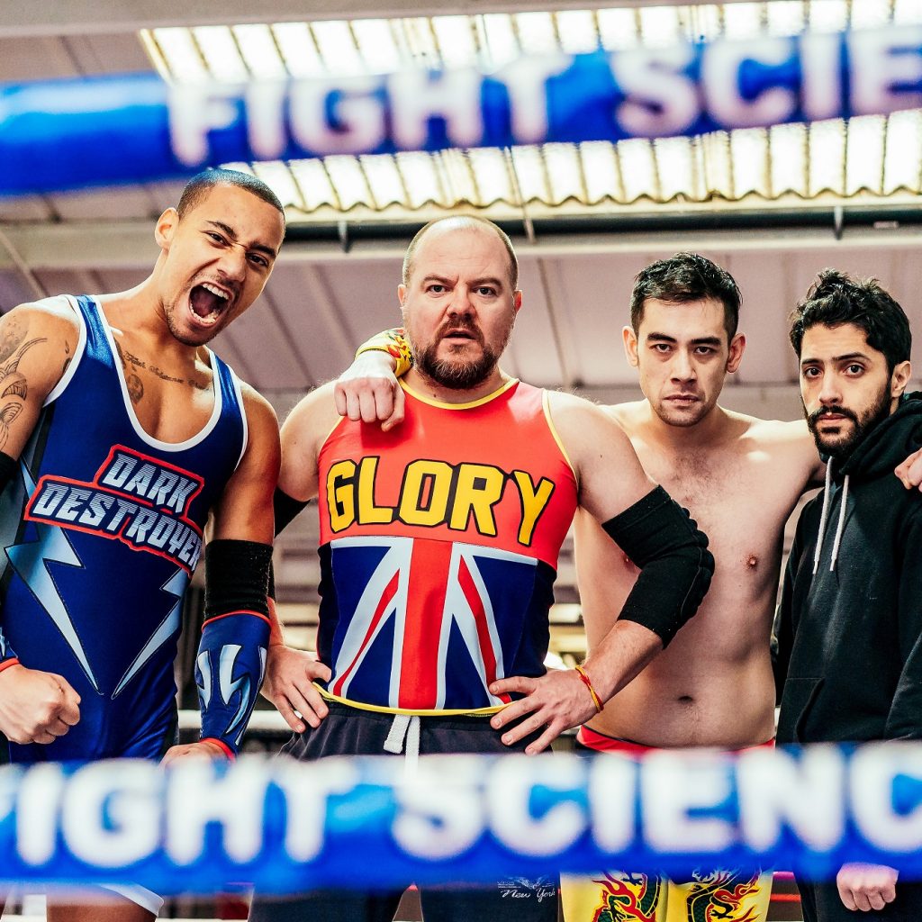 Four men look out at the camera through the bright blue ropes of a wrestling ring.