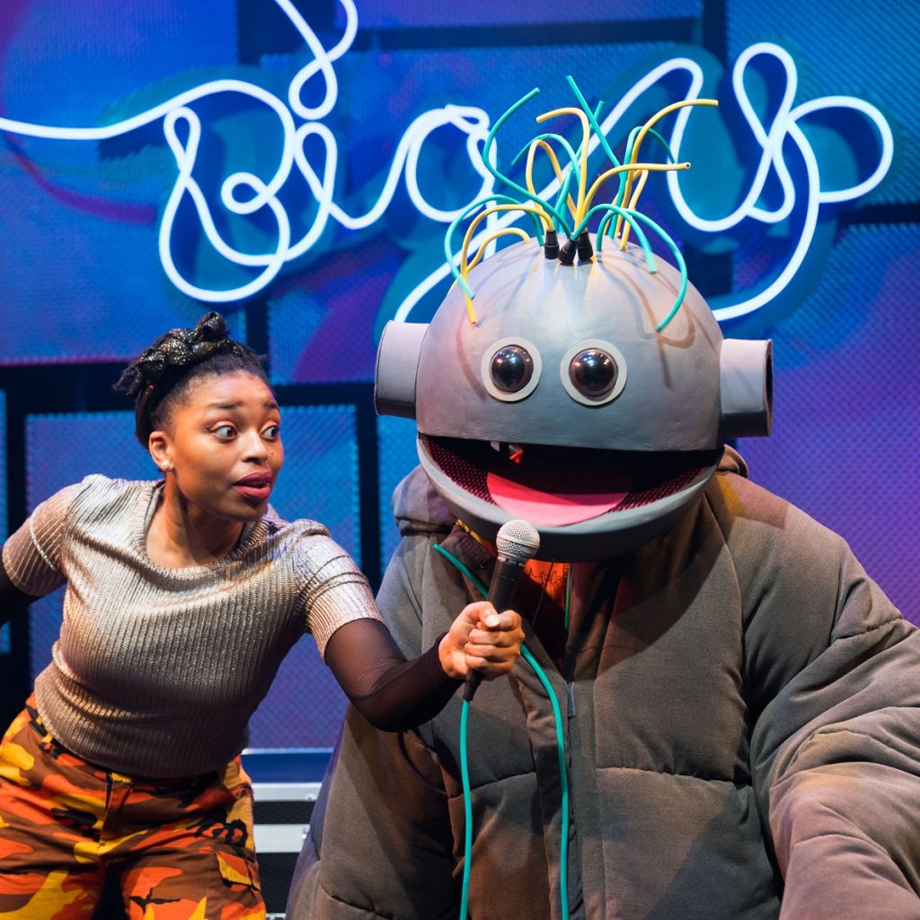 A female actor holds a microphone to the mouth of a life-size puppet