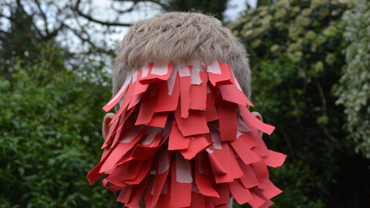 A man with strips of paper obscuring his face looks to camera.