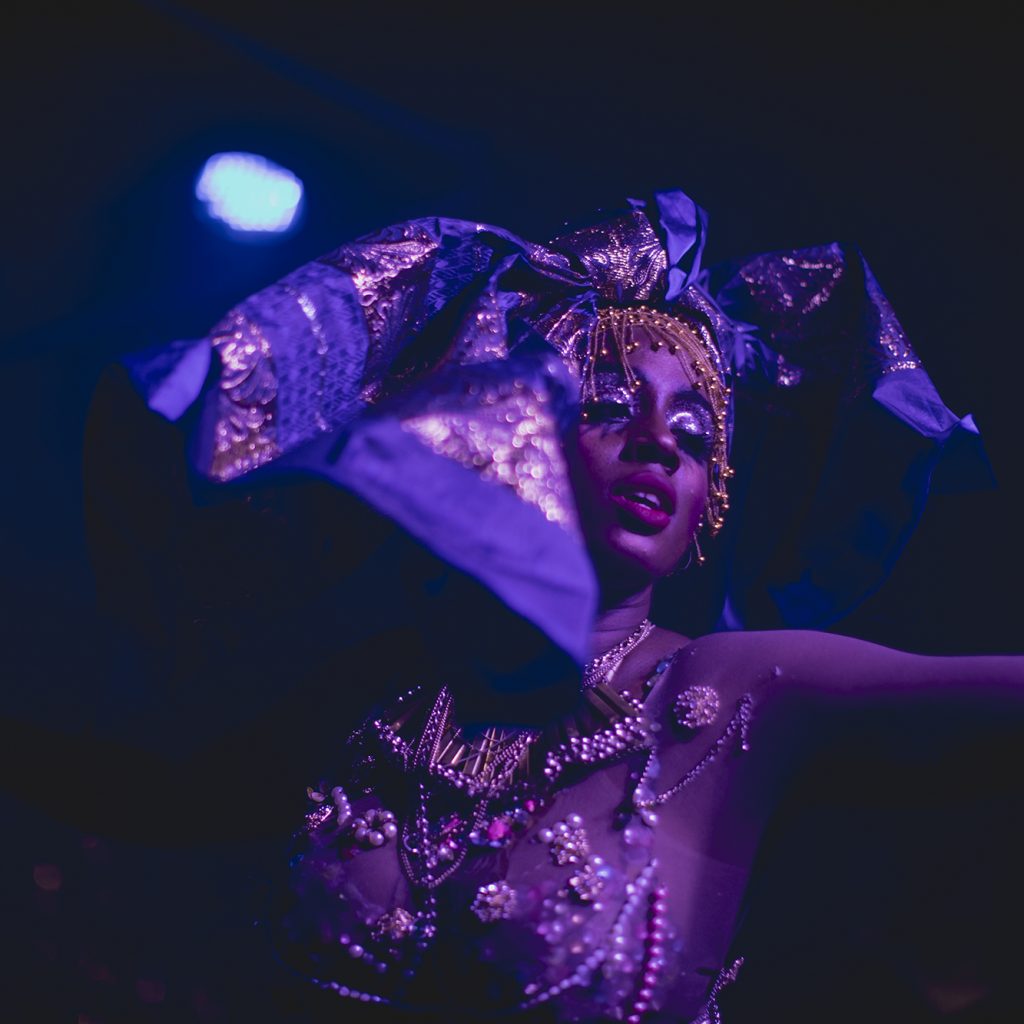 A black woman wearing a large colourful head tie and a jeweled outfit poses with her arms out.