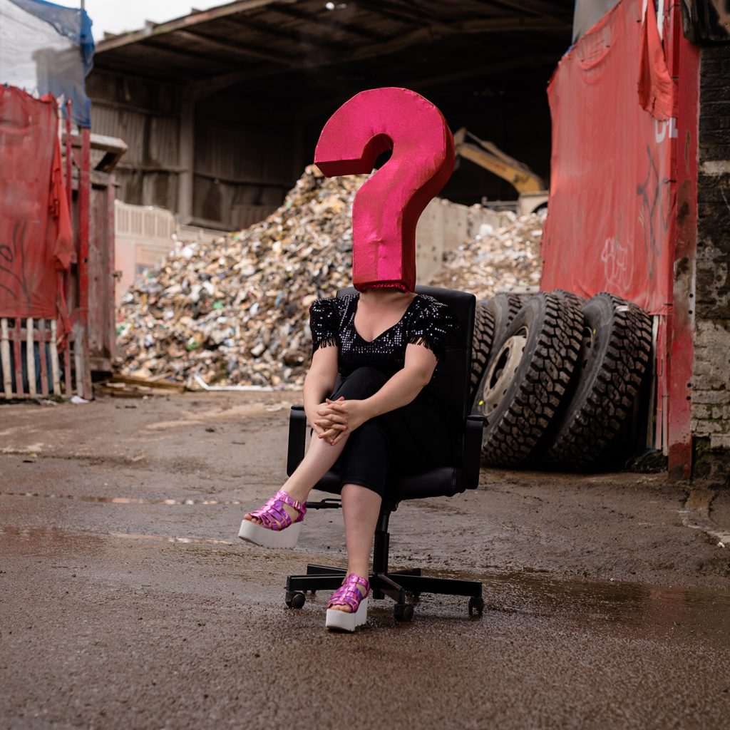 A woman sat on a seat with her legs crossed with a large pink question mark on her head covering her face.