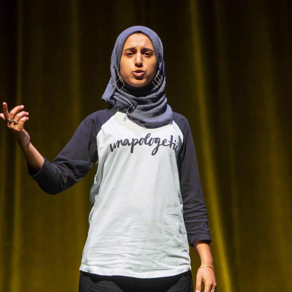 A young woman in a headscarf speaks from a stage.