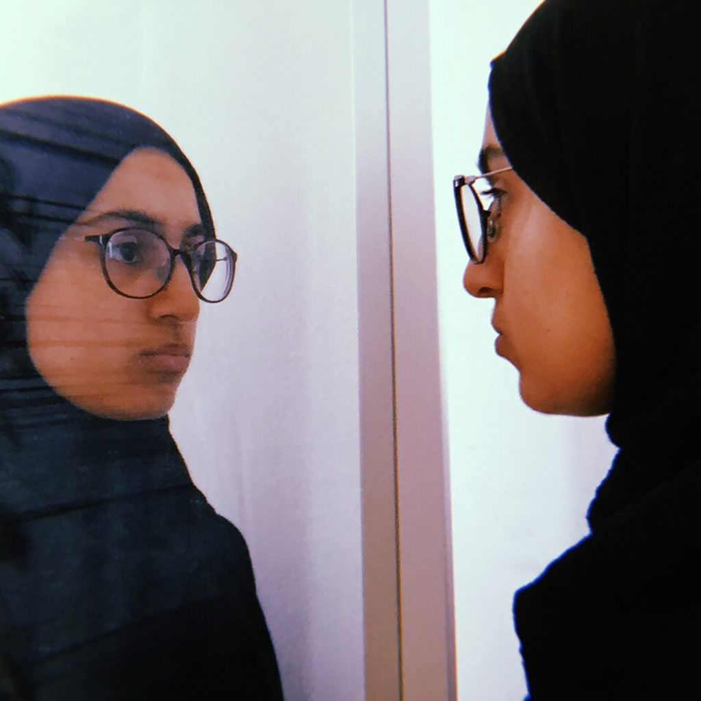 A young woman in a headscarf looks at herself in the mirror.