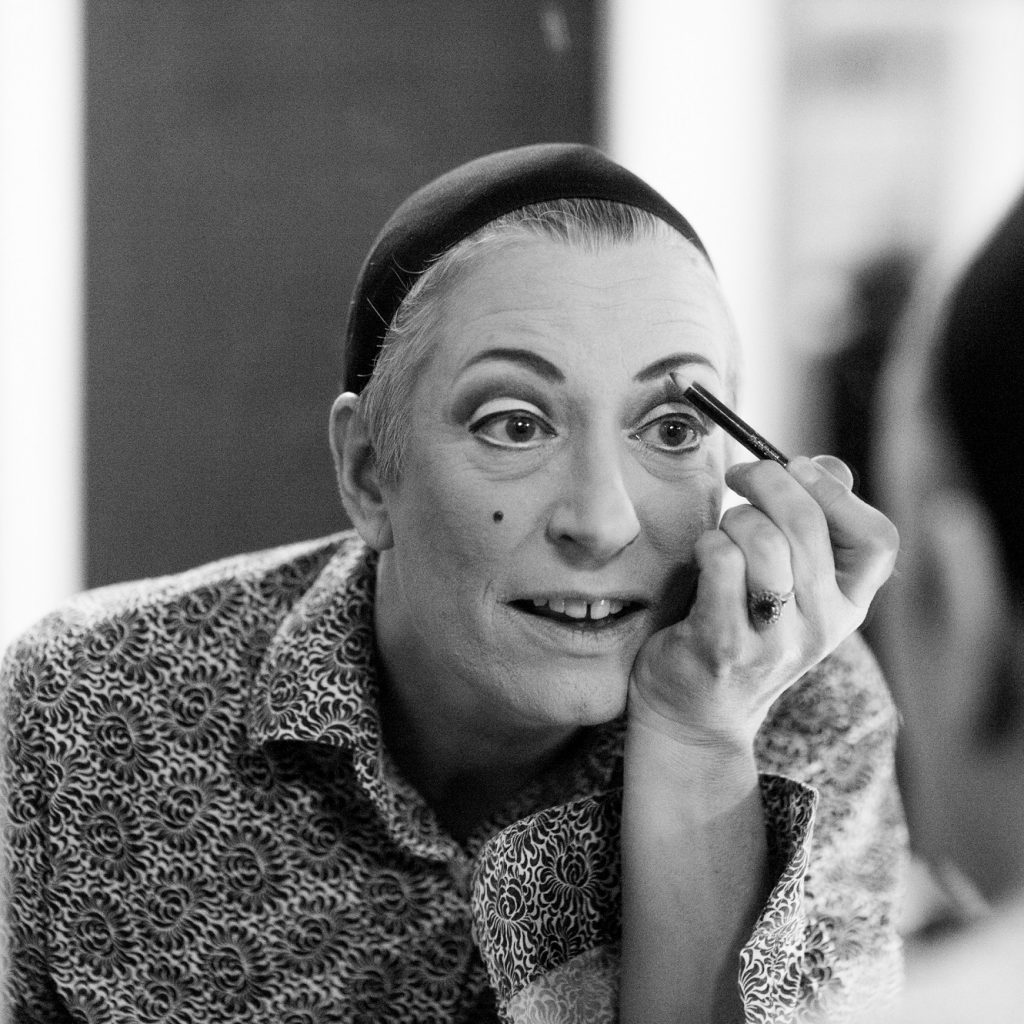 Performer pictured in black and white in a ,mirror, applying make-up.