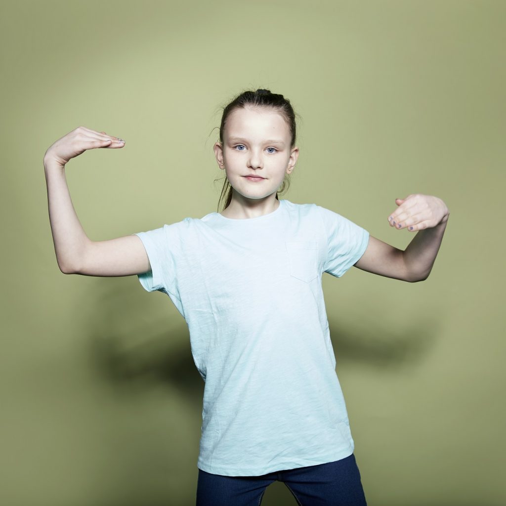 A girl in a pale blue T-shirt poses in a dance move looking confidently at the camera.