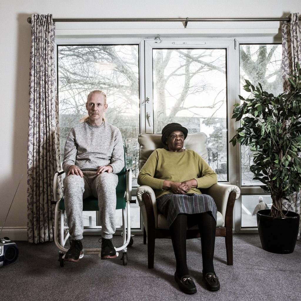 Image of two people sitting on chairs, staring straight ahead in a care home.