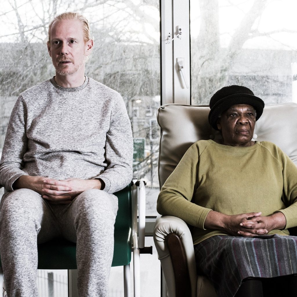 Image of two people sitting on chairs, staring straight ahead in a care home.