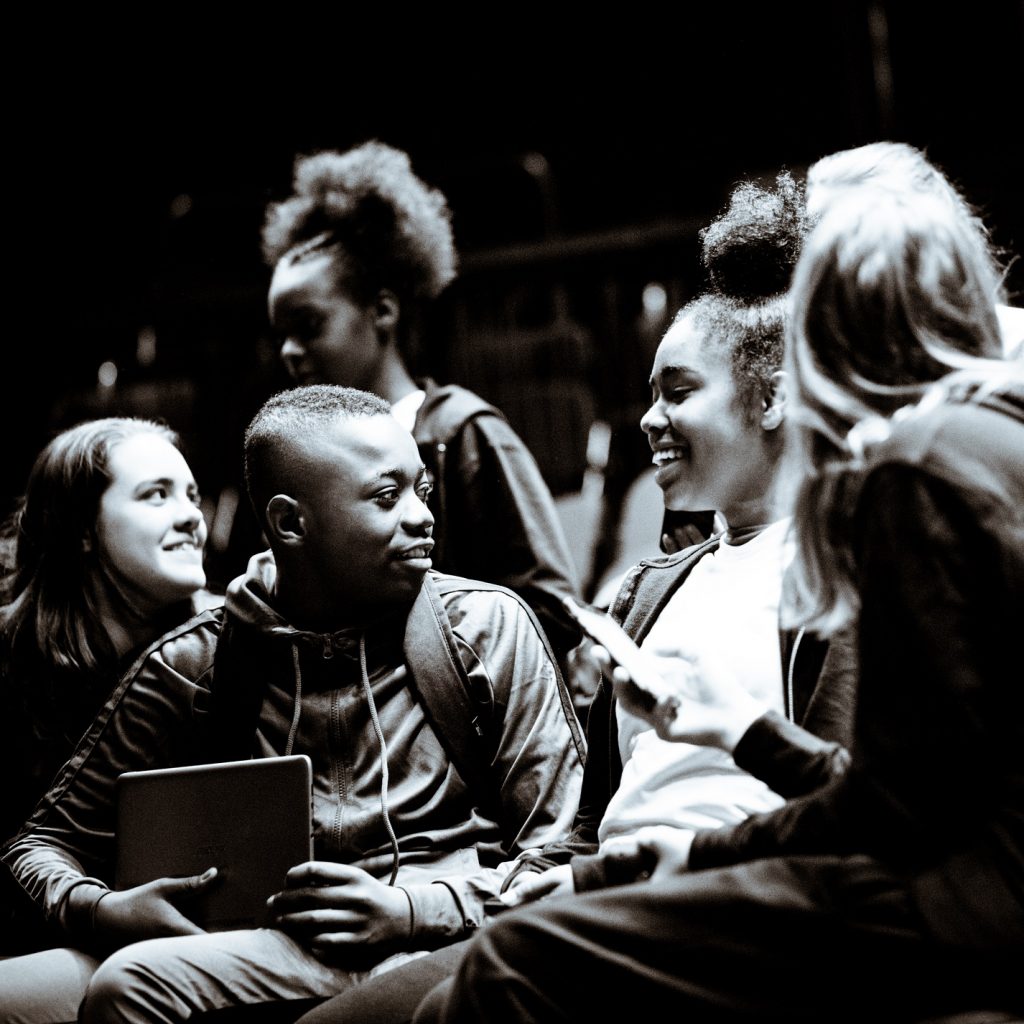 Black and white image of a group of children sitting in an audience, talking and smiling.