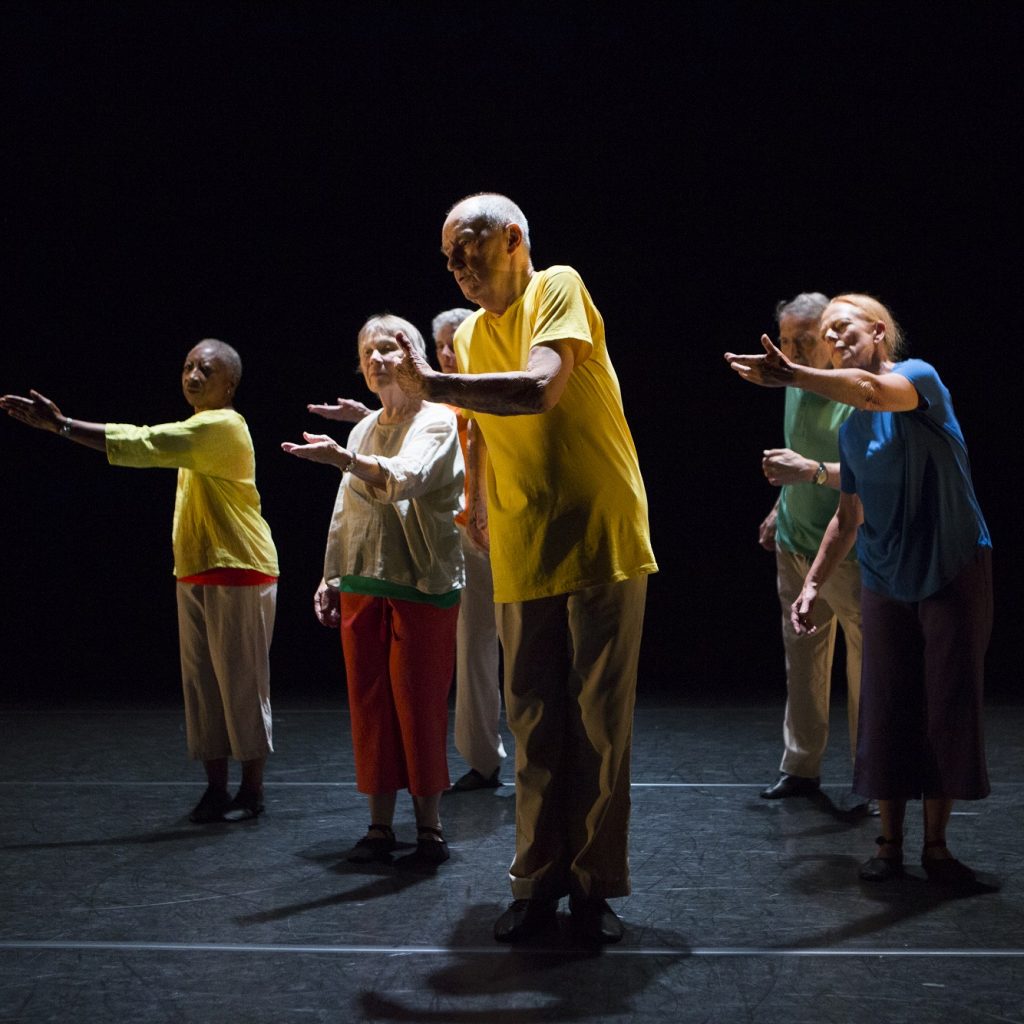 A group of people dancing on a dark stage, with one arm outstretched.