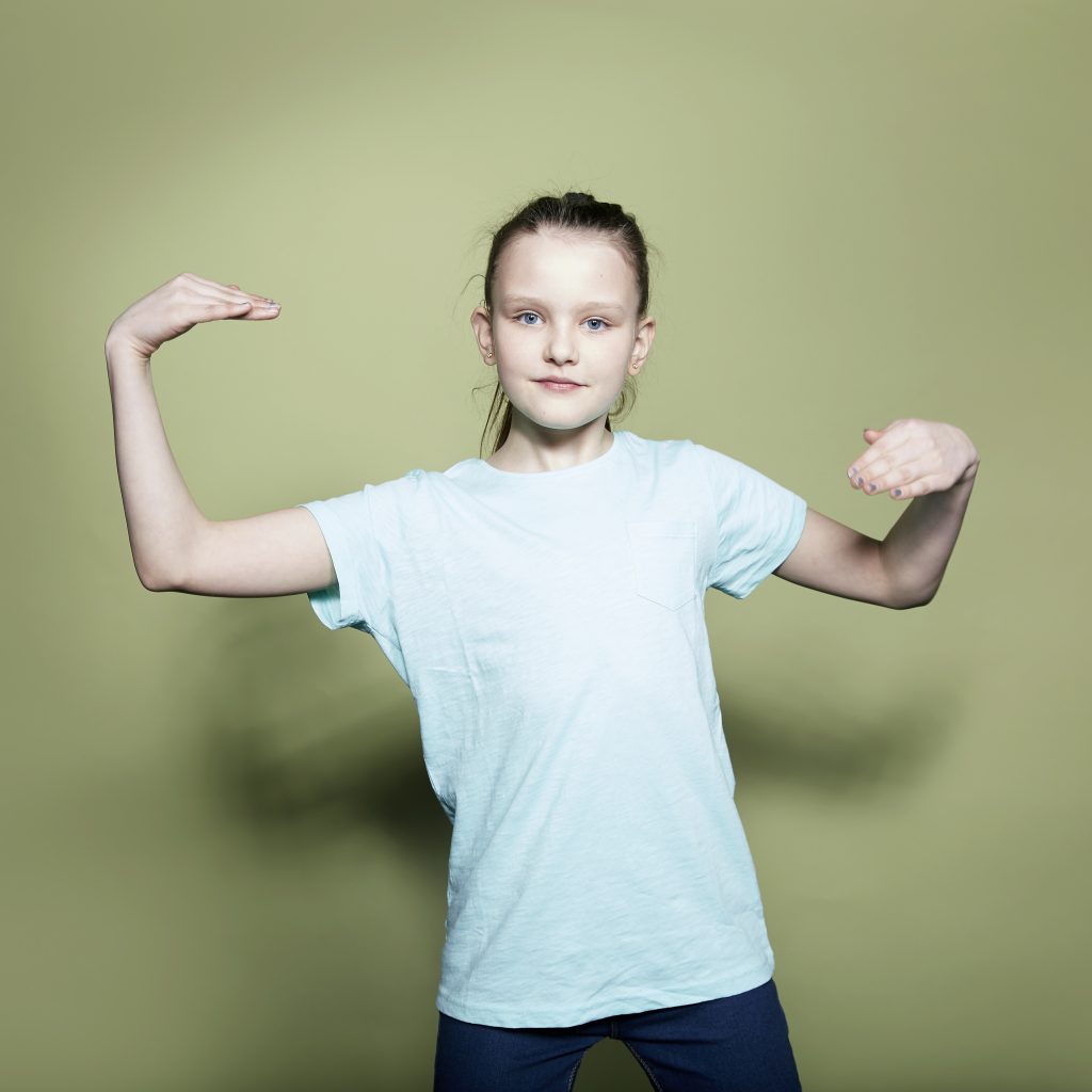 Image of a young girl doing a dance move, standing against a green background.