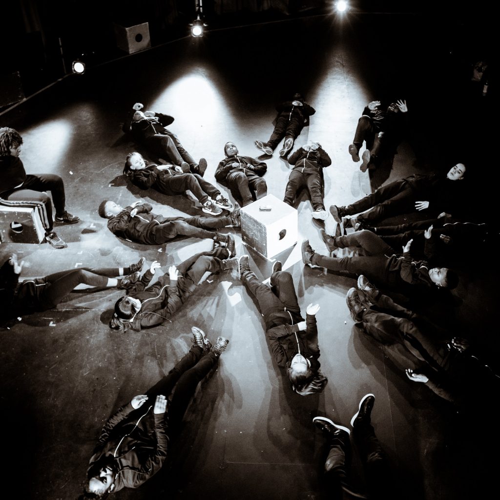 Black and white image of performers on a stage from a birds-eye view. They are all lying on their backs on the floor in a circle.