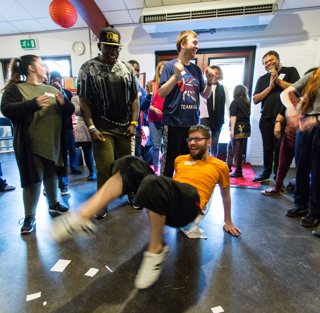 A group of young people in a circle. A young man wearing a yellow shirt is in the middle on the floor break dancing.