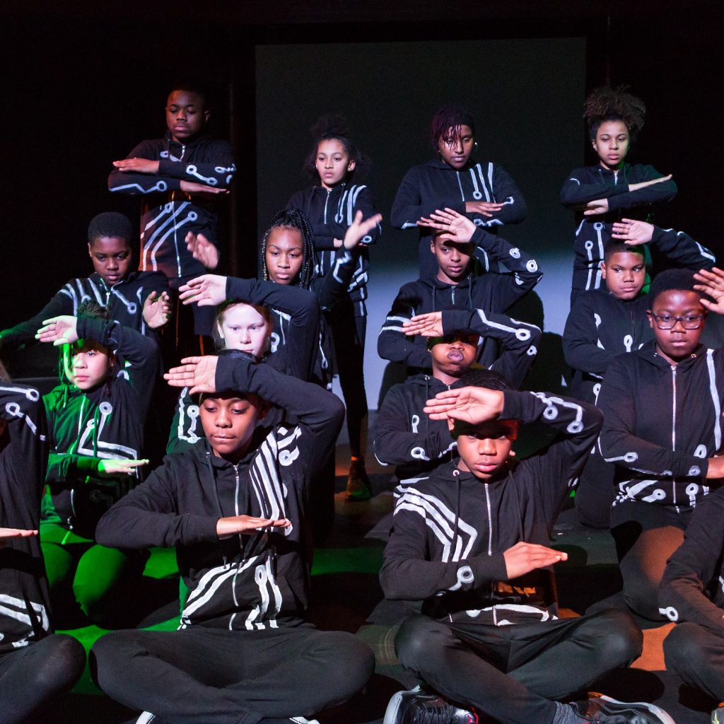 Image of a group of people sitting on stage, doing a dance move with their arms