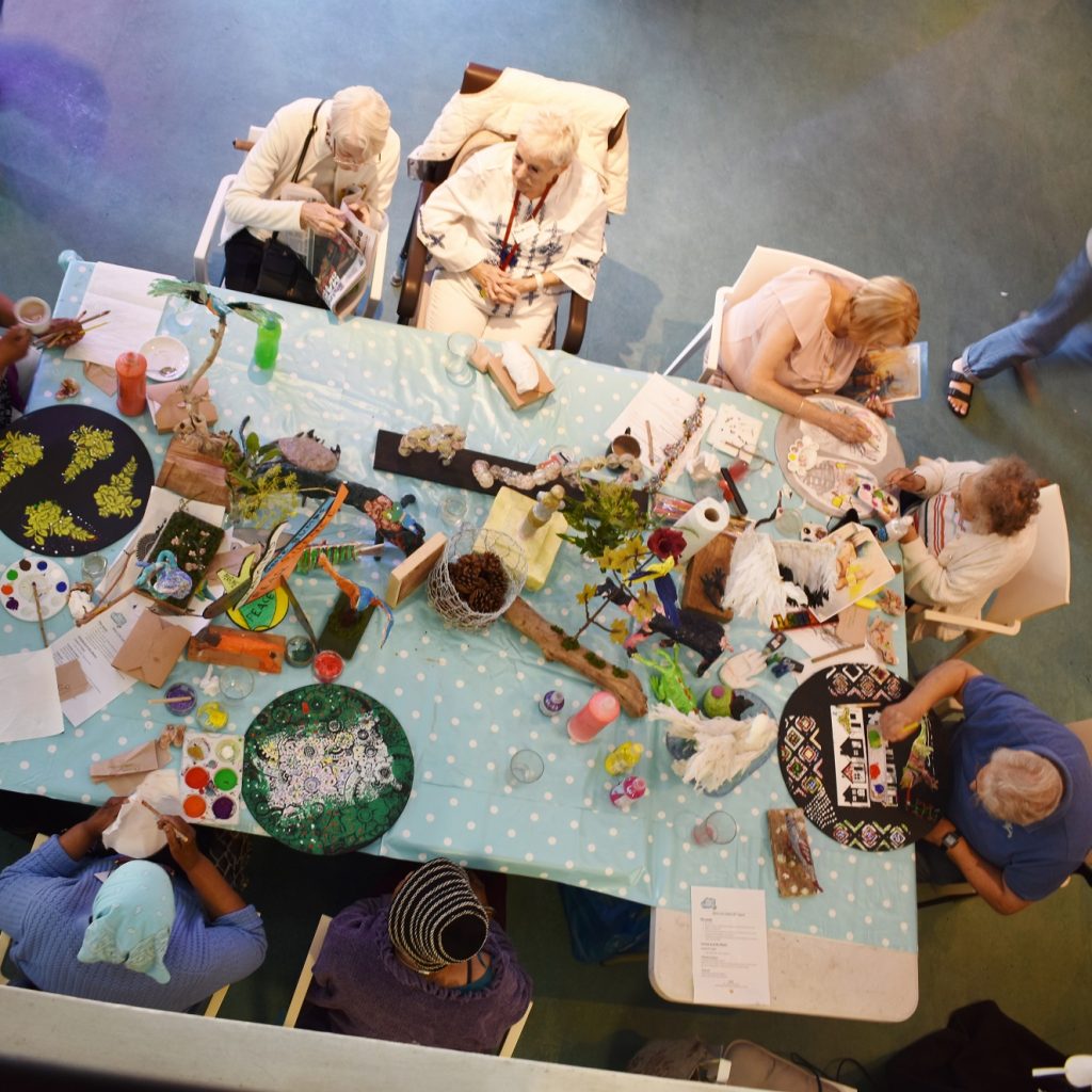 Table full of art work and older artists, viewed from above.