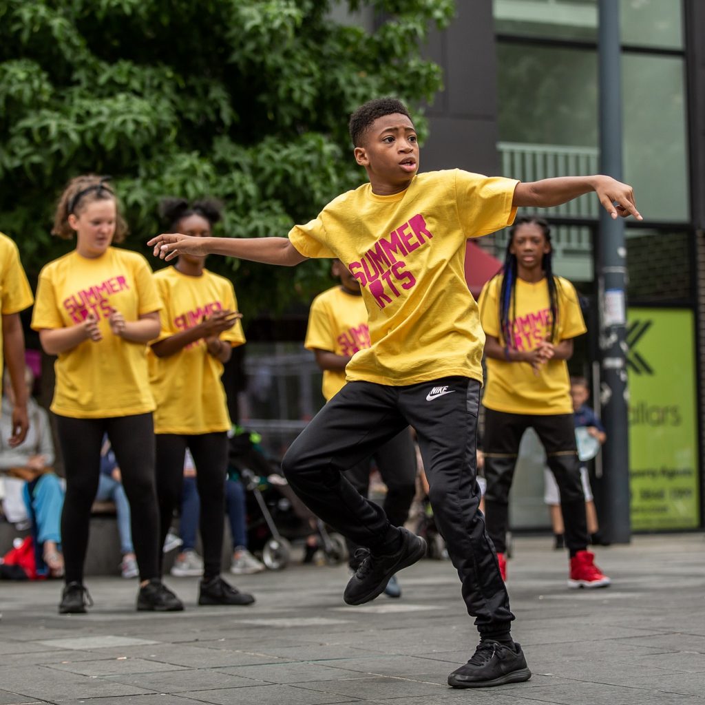 A young Summer Arts participant hip hop dancing on Giffin Square. Behind watching are other participants