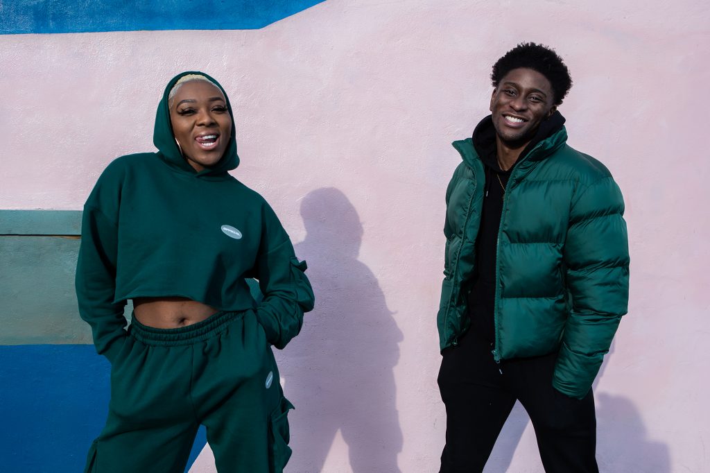 A lady with a green hoodie and traksuit bottoms sticking here tongues out and a man in a green puffer jacket smiling in front of a pink wall