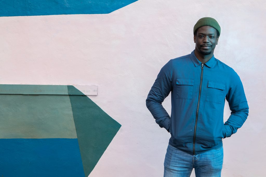 A man stood in front of a pink wall wearing a green hat and blue jacket, smiling with his hands in his pockets