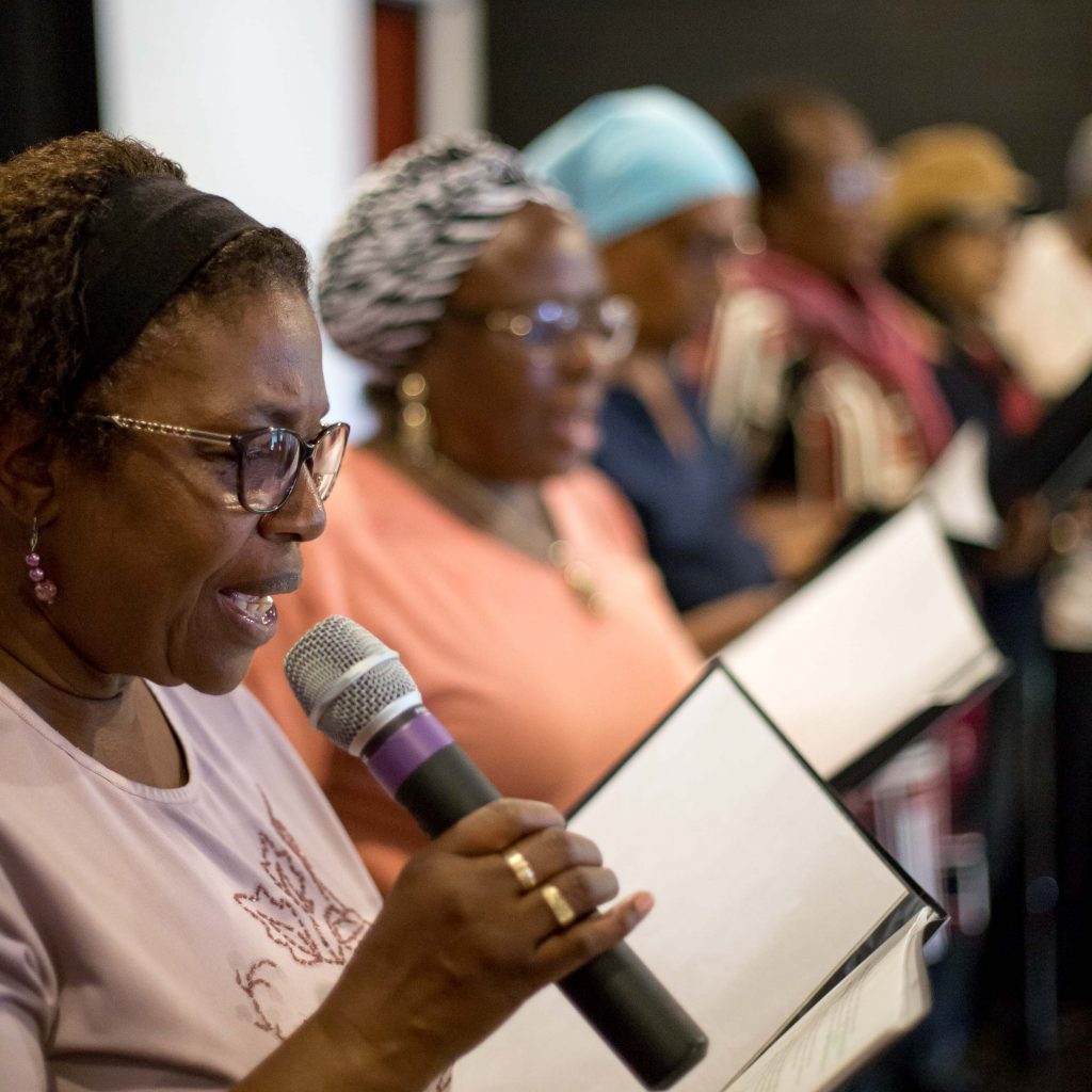 Meet Me member, a black woman wearing a pale pink shirt, singing in the choir with other members, holding a microphone and the lyrics book