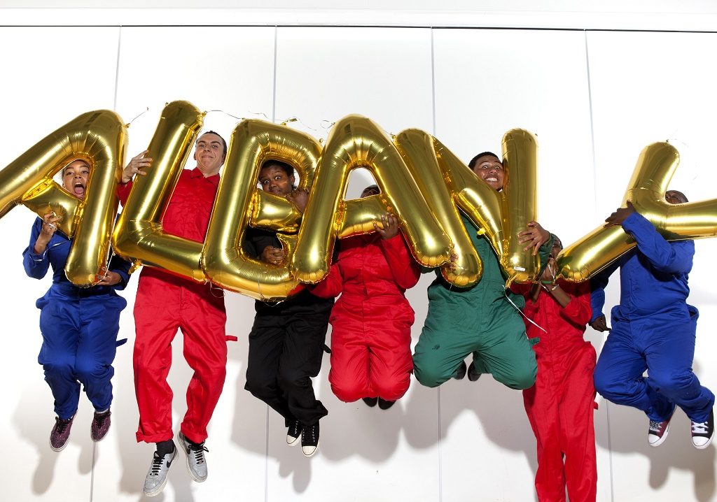 Seven young people in boiler suits jumping, all mid-air, holding letter-shapred balloons that spell 'ALBANY.'