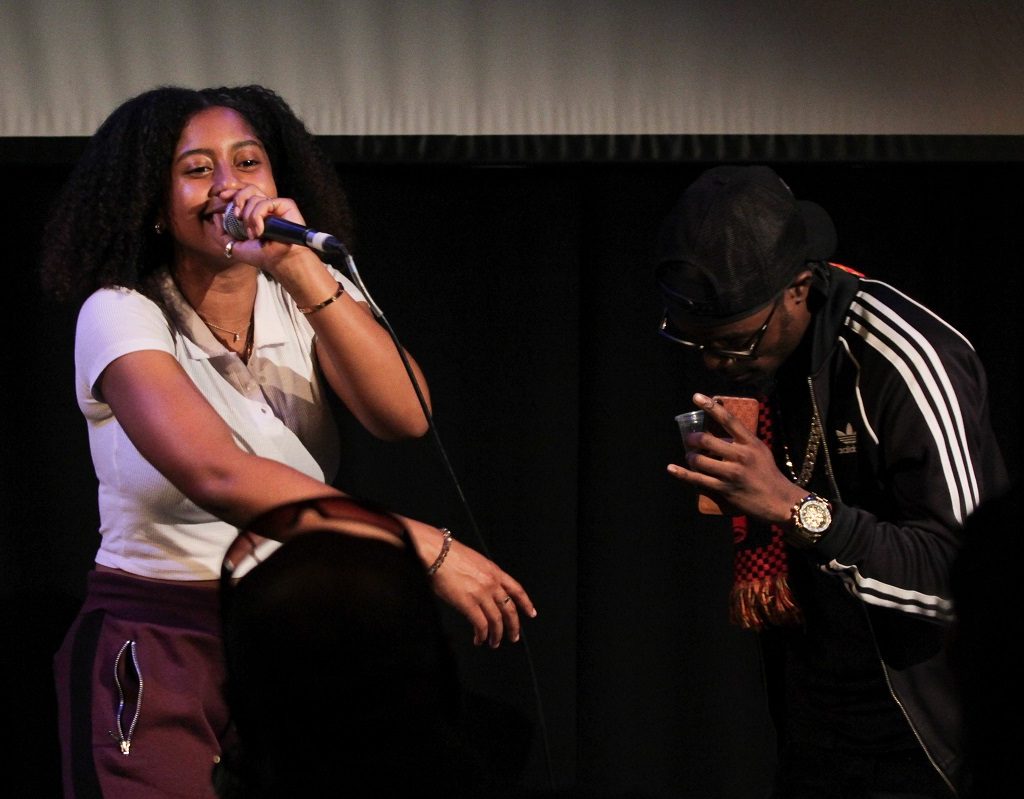 A young woman with curly hair is wearing a white t-shirt and holding microphone. She's standing next to a young man dressed in black with his head down.