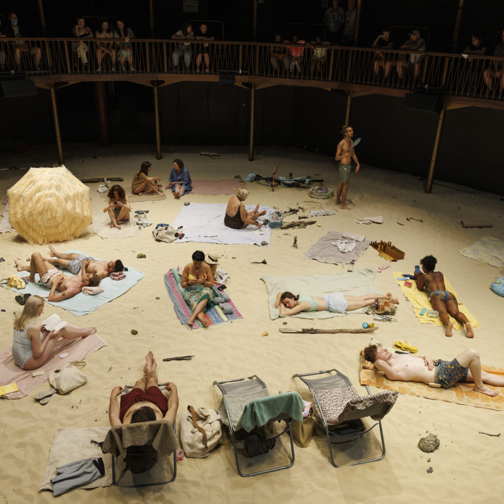 The Albany auditorium filled with sand. Beachgoers sunbathe and there are towels, umbrellas and sun loungers dotted about.