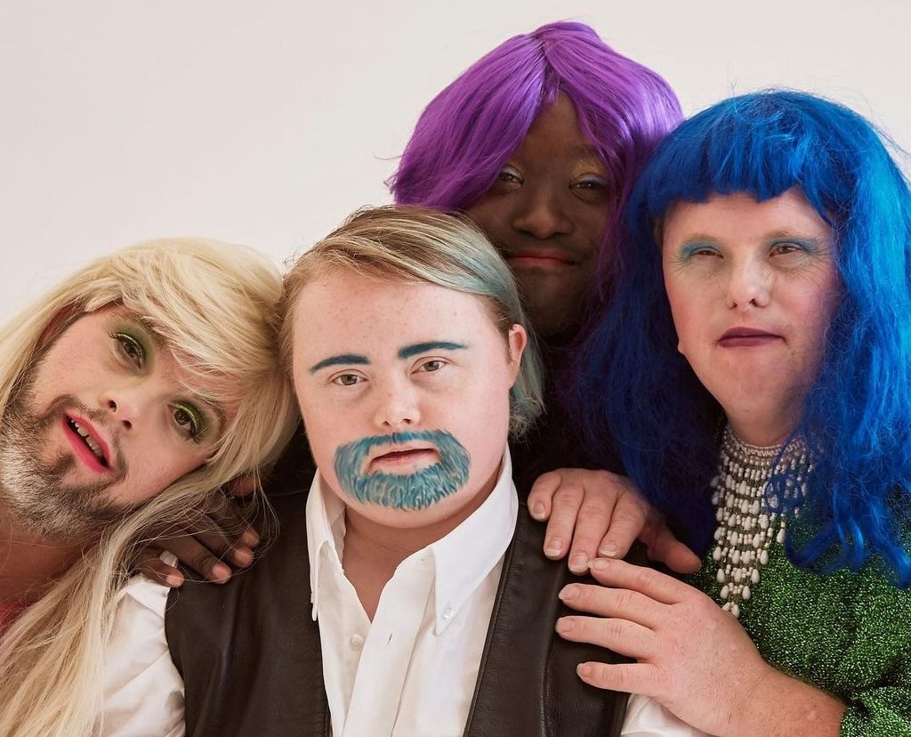 4 people in drag pose for a photo together, three wear bright wigs, the fourth has a blue painted moustache and eyebrows.
