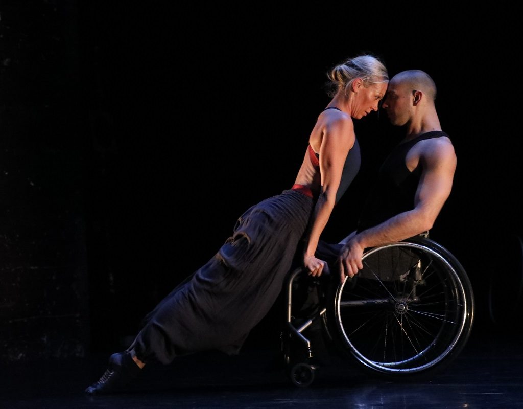 Two dancers perform together. One of them is using a wheelchair. They are close together with their foreheads touching.