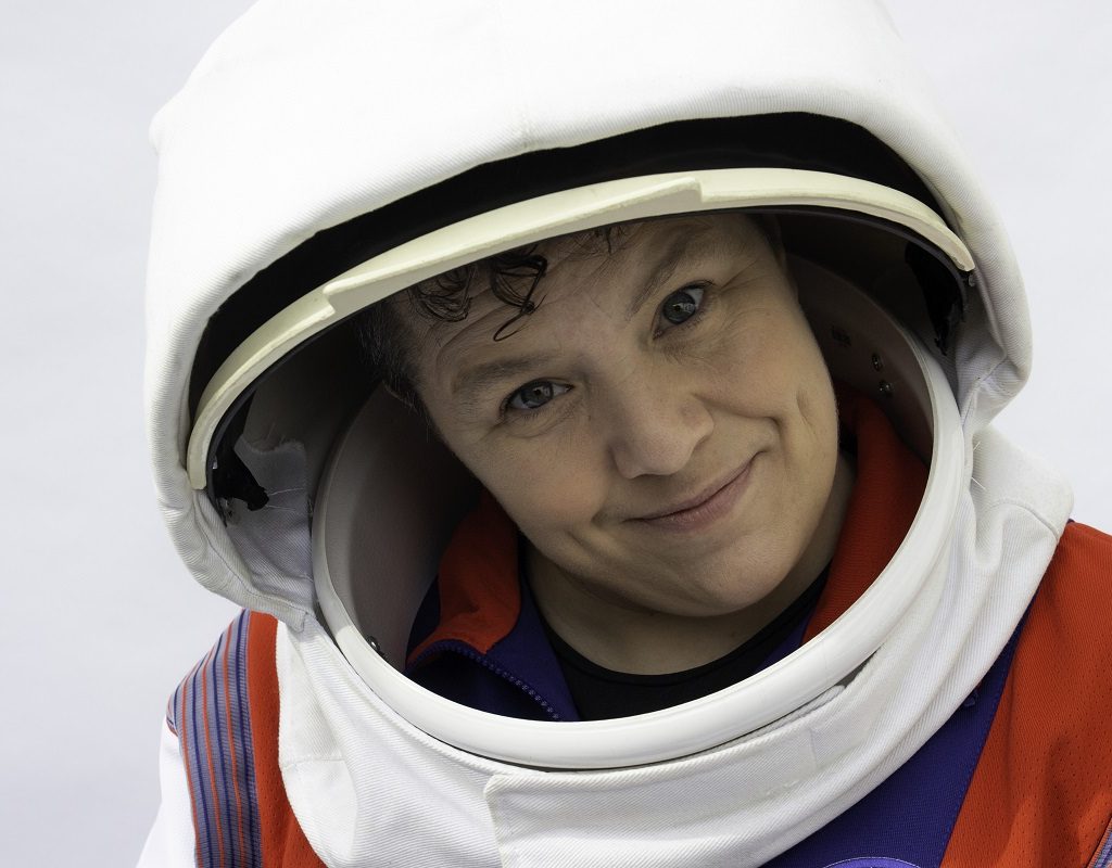 A person (Jess Thom) sits in a wheelchair wearing an astronaut's suit. She smiles, her arms outstretched towards us.