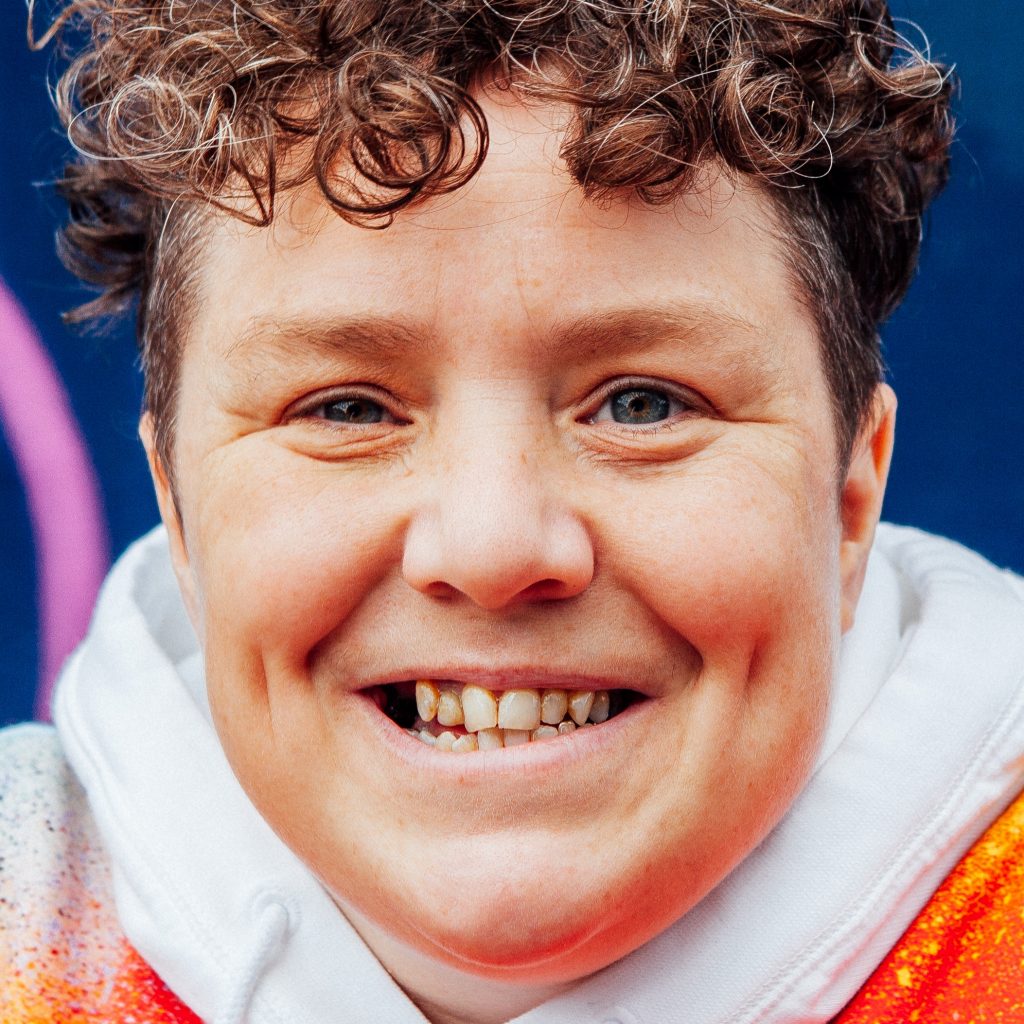 A person with short curly hair looks at the camera with a cheeky smile. They are wearing an orange hooded sweatshirt.