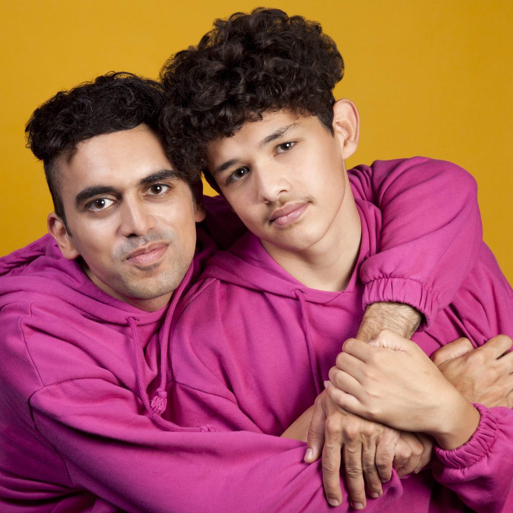 A man and boy wear matching pink jumpsuits and look towards the camera. The man has his arms around the boy. The background is a deep yellow.