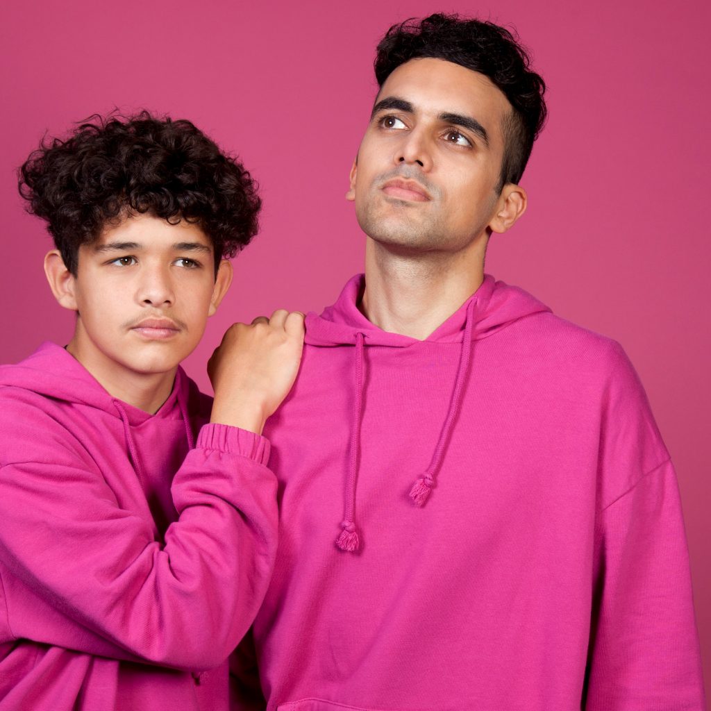 From left to right: A young man with curly hair wearing a pink sweatshirt with a hood, and is leans his left hand on another mans shoulder. This man is also wearing a pink sweatshirt with a hood.