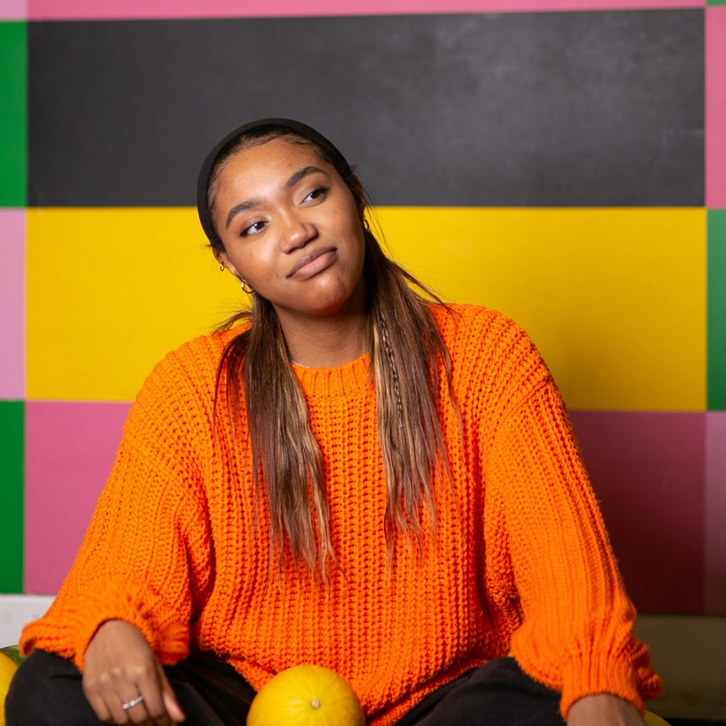 A young person wearing an orange jumper sits in front of a background that has blocks of different colours.