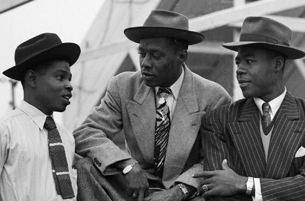 An image showing The Windrush generation. Three black men wearing suits and ties and hats converse with each other.