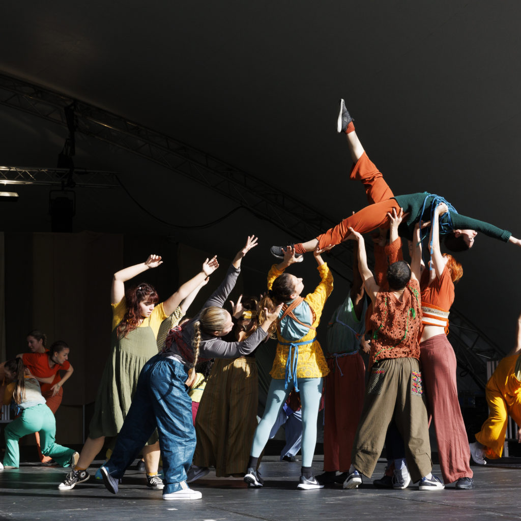 A group of people wearing bright colours dance on an outdoor stage.