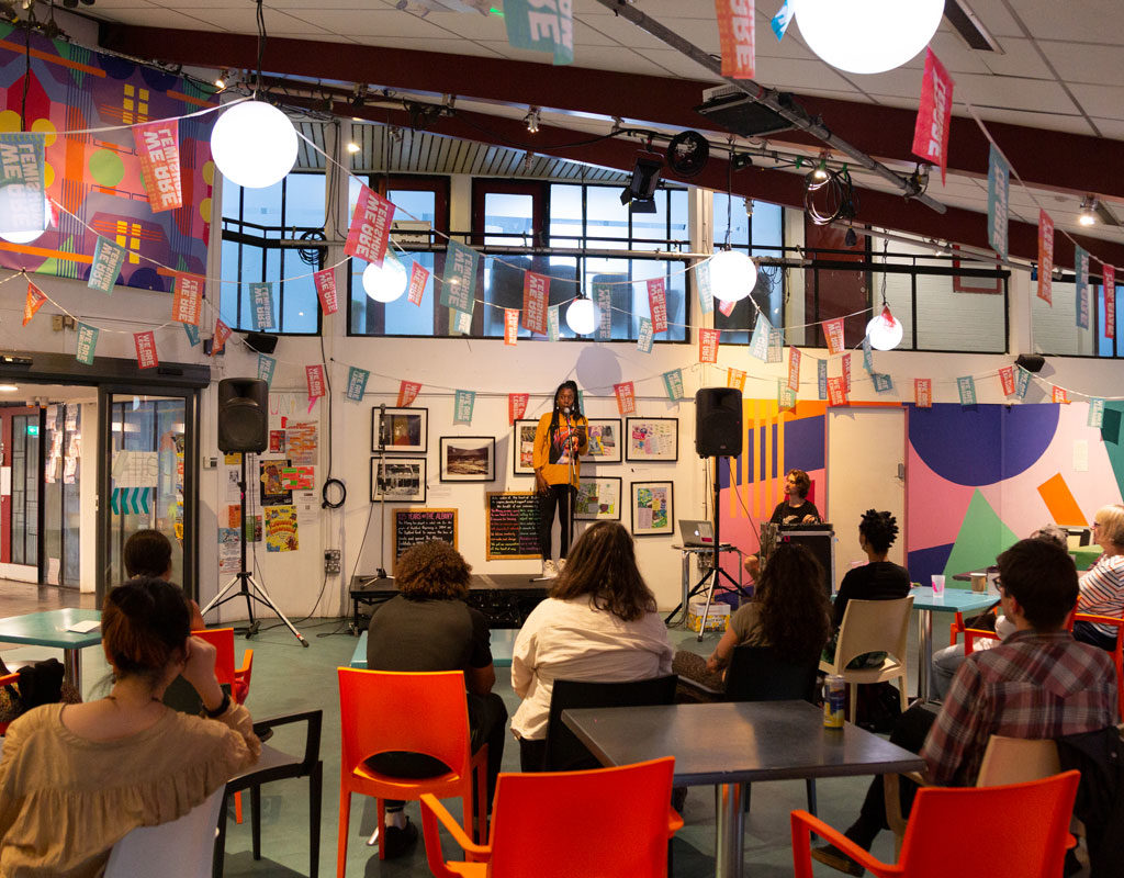 Pizza and Pitches event at the Albany CaffA. One of the contestants, a Black woman wearing black jeans and an orange long-sleeved top, talks to a microphone. Audience members watch her.
