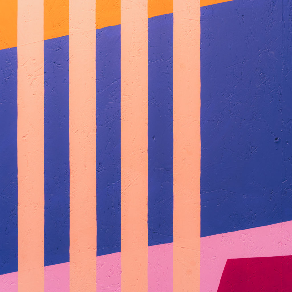 The mural wall at the Albany CaffA. Five vertical peach lines, a diagonal blue parallelogram layered underneath, the bottom panel baby pink with a fuscia rhombus at the bottom over the pink. Top corner an orange triangle.