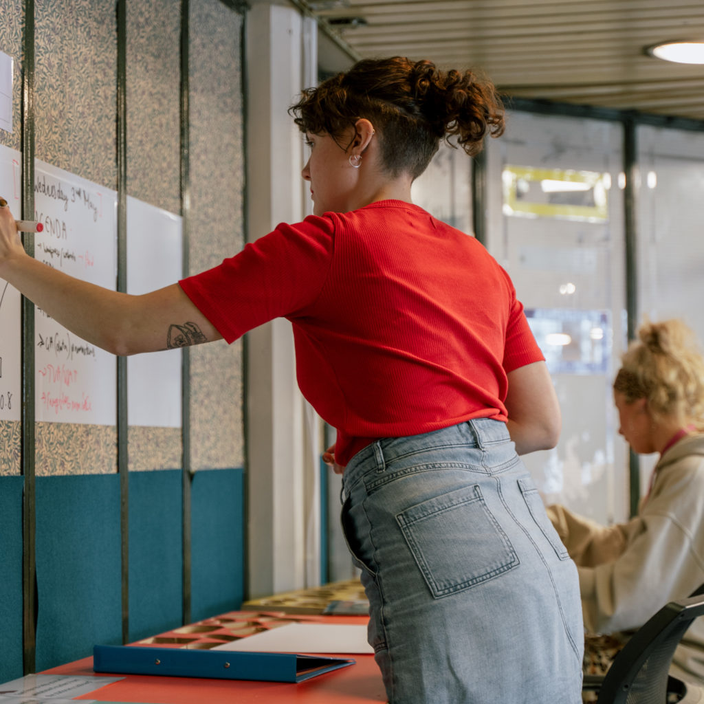 White woman with a red t-shirt and denim skirt using the whiteboard in the creative workspace. Sat next to her is a white woman with bleach blond hair wearing a light grey jumper collaging at one of the desks